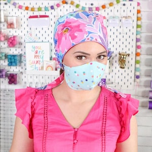 Puede incluir: Una mujer con una camisa rosa con un gorro quirúrgico de estampado floral y una mascarilla azul y blanca con lunares. La mascarilla tiene un diseño de corbata.
