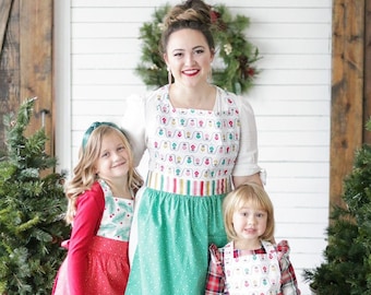 May include: A woman and two girls wearing red and green Christmas-themed aprons with a white background and a pattern of Christmas lights. The aprons have a green polka dot skirt and a white bodice with a red and white striped band.