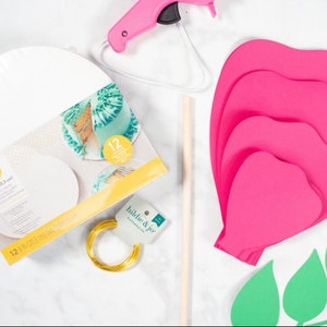 May include: A set of craft supplies for making a paper flower. The supplies include a hot glue gun, a wooden dowel, a roll of gold wire, a package of 8 inch round cake boards, and pink and green paper cutouts of flower petals and leaves.