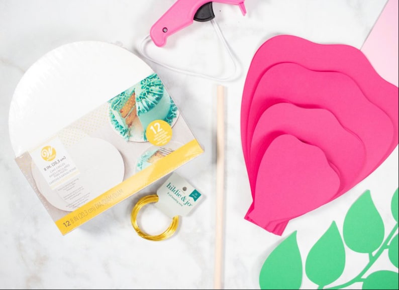 May include: A set of craft supplies for making a paper flower. The supplies include a hot glue gun, a wooden dowel, a roll of gold wire, a package of 8 inch round cake boards, and pink and green paper cutouts of flower petals and leaves.