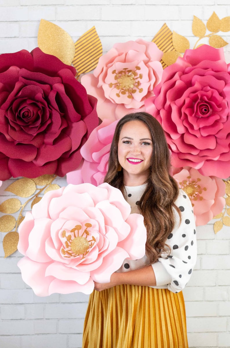 May include: A woman is holding a large pink paper flower in front of a wall decorated with several large paper flowers in shades of pink and burgundy. The flowers are all made of paper and have a layered, ruffled appearance.