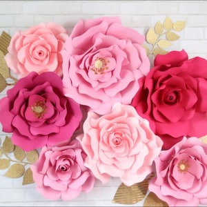 May include: A set of seven large paper flowers in shades of pink and red. The flowers are arranged on a white brick wall background. The flowers are made of paper and have a layered, ruffled appearance.