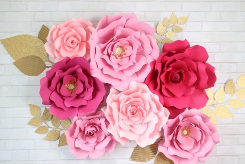 May include: A set of seven large paper flowers in shades of pink and red. The flowers are arranged on a white brick wall background. The flowers are made of paper and have a layered, ruffled appearance.