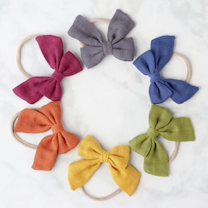 May include: Set of six fabric hair bows in various colors, including mustard yellow, burnt orange, burgundy, dark blue, and gray. Each bow is tied with a nude elastic headband.