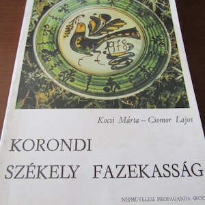 May include: A book cover featuring a green and white ceramic plate with a bird design. The title of the book is "Korondi Székely Fazekaság" and the author is "Kocsi Márta - Csomor Lajos".