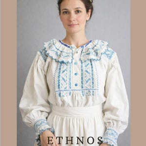 Könnte beinhalten: Eine Frau in einem weißen Volkskleid mit blauer Stickerei und Rüschenkragen. Das Kleid hat lange Ärmel und gehört zur "ETHNOS"-Kollektion, beschrieben als Volkskleidung aus Transsilvanien von E.D. Stupar.