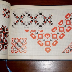 Könnte beinhalten: Aufgeschlagenes Buch mit verschiedenen Kreuzstichmustern in Rot, Schwarz und Weiß. Die Designs sind geometrisch, mit Quadraten, Rauten und blumenartigen Motiven. Die Seiten sind kariert, mit einem blauen Lesezeichen.