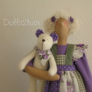 Op de afbeelding: Een paars-groene jurk met witte stippen en een ruitpatroon. De jurk heeft een witte ruche en een witte teddybeer met paarse oren en een bloemen sjaal.
