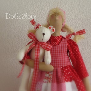 Op de afbeelding: Een rood-wit geruit jurkje met een rood gestippeld bovenstuk en een wit-roze patchwork rok. De pop houdt een witte teddybeer vast met een rood-wit geruite strik.