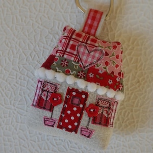 Könnte beinhalten: Ein roter und weißer Patchwork-Schlüsselanhänger in Form eines Hauses mit einem Herz auf der Vorderseite. Der Schlüsselanhänger hat einen silbernen herzförmigen Ring.