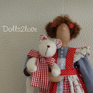 Puede incluir: Una muñeca hecha a mano con cabello castaño y un vestido de cuadros rojos y blancos sostiene un animal de peluche blanco. La muñeca lleva un delantal de estampado floral azul y blanco. La muñeca sostiene un animal de peluche blanco con un lazo de cuadros rojos y blancos.