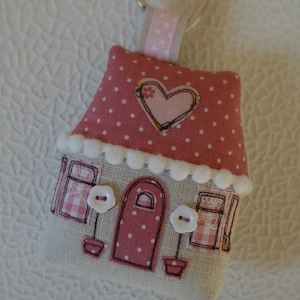 Op de afbeelding: Een roze en witte huisvormige sleutelhanger met een hart op het dak en witte knopen voor ogen. De sleutelhanger heeft een zilveren ring om aan sleutels te bevestigen.