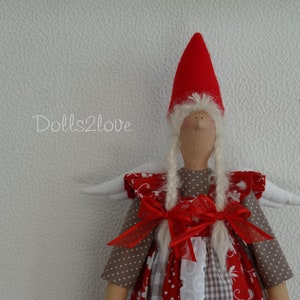 Könnte beinhalten: Eine handgemachte Puppe mit einem roten Hut, weißen Flügeln und einem rot-weißen Kleid. Die Puppe hat langes weißes Haar und einen süßen Ausdruck.