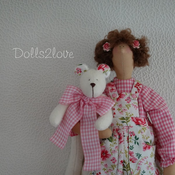 La poupée Tilda Sammy-Jo porte une belle salopette blanche avec un imprimé de roses accompagnée de son petit ours en peluche.