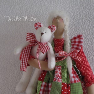 Peut inclure: Une poupée de chiffon blanche avec de longs cheveux blancs portant une robe en patchwork rouge et verte avec un ours en peluche blanc dans ses bras. La poupée tient l'ours en peluche avec un ruban à carreaux rouge et blanc.