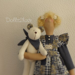 Peut inclure: Une poupée faite main avec des cheveux blonds et une robe en patchwork bleu et marron tient un ours en peluche blanc avec une écharpe à pois bleus et blancs. La poupée a des fleurs blanches dans les cheveux. L'arrière-plan est un mur texturé blanc avec le texte "Dolls2love" dans le coin supérieur gauche.