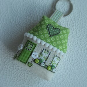 May include: A green and white fabric house-shaped keychain with a white heart and white buttons. The keychain has a silver ring.