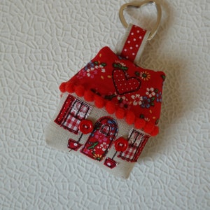 Puede incluir: Un llavero de tela rojo y blanco con forma de casa con un corazón en el techo y botones rojos para los ojos. El llavero está unido a un anillo de corazón plateado.