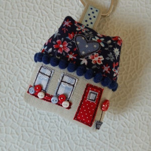 May include: A blue and white fabric house-shaped keychain with a red door and a heart on the roof. The keychain has a silver heart-shaped ring.