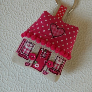 Può includere: Un portachiavi a forma di casa con pois bianchi su sfondo rosa. La casa ha un cuore rosso e un bordo bianco. La porta è rossa con un fiore bianco e due bottoni bianchi. Il portachiavi ha un anello a forma di cuore argentato.