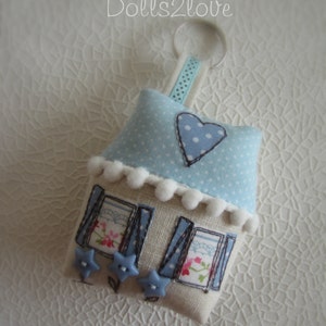 May include: A blue and white fabric house-shaped keychain with a white heart and blue polka dots. The house has white trim and blue windows with floral designs. The keychain has a blue ribbon and a silver ring.
