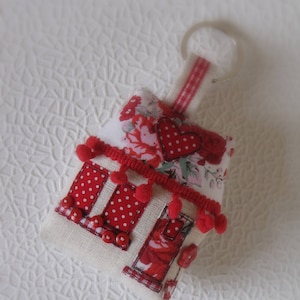 Può includere: Un portachiavi in tessuto bianco a forma di casa con dettagli in tessuto a pois rossi e bianchi, pompon rossi e un cuore rosso. Il portachiavi ha un anello argentato.