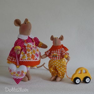 Puede incluir: Dos muñecos de ratón de tela. El ratón más grande lleva un vestido rosa de lunares con un delantal floral y sostiene un adorno en forma de corazón. El más pequeño lleva una camisa de cuadros naranja y pantalones amarillos de lunares, tirando de un coche de juguete de madera.