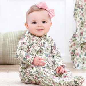 May include: A smiling baby wearing a light green footed onesie with a floral and gardening tool pattern. The onesie has long sleeves and a ruffled collar. A pink bow adorns the baby's head. The baby is sitting on a white rug.
