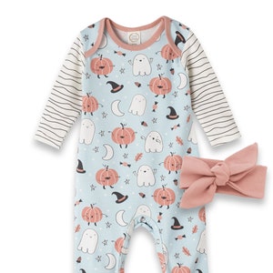 May include: A baby onesie with a blue background and a pattern of orange pumpkins, white ghosts, and stars. The onesie has long sleeves with a white and black striped pattern. The onesie has pink trim around the neck, sleeves, and legs. A pink bow headband is included.