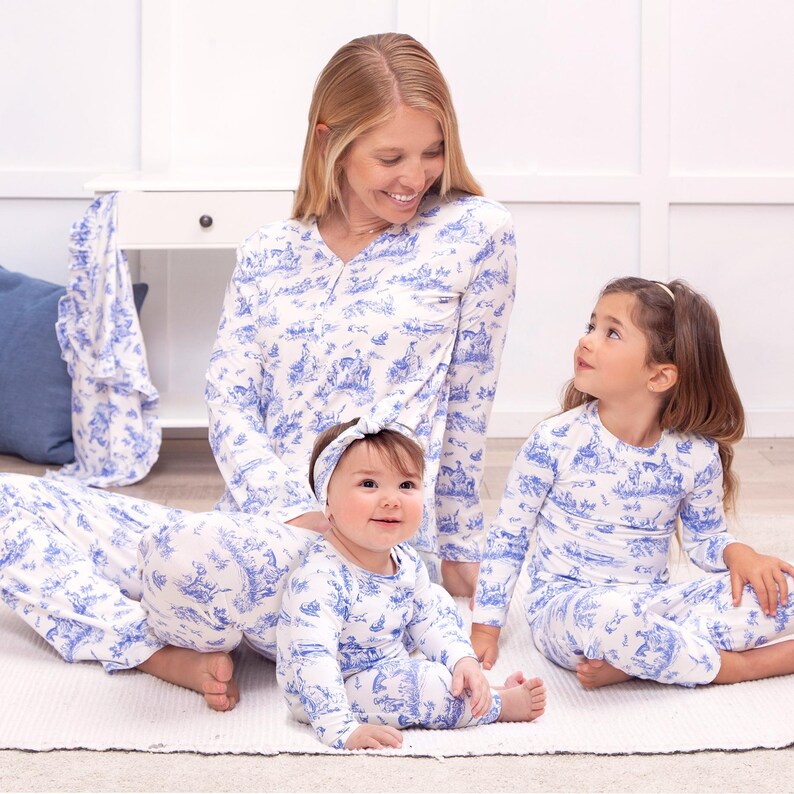 May include: Matching family pyjama sets in a white and blue toile print. The sets include long-sleeved tops and trousers for adults and children, and a headband for the baby. The family is sitting on a white rug.
