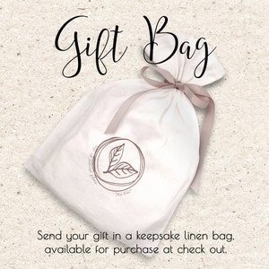 May include: A white linen gift bag tied with a light brown ribbon. The bag features a circular logo with two leaves and the text "Tella Babe". The words "Gift Bag" are written in cursive at the top. The text "Send your gift in a keepsake linen bag" is at the bottom.