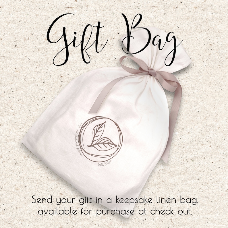 May include: A white linen gift bag tied with a light brown ribbon. The bag features a circular logo with two leaves and the text "Tella Babe". The words "Gift Bag" are written in cursive at the top. The text "Send your gift in a keepsake linen bag" is at the bottom.