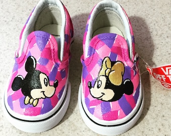 May include: A pair of pink and purple Vans slip-on shoes with a Mickey Mouse and Minnie Mouse design. The shoes are painted with a pink and purple checkered pattern.