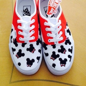 Puede incluir: Un par de zapatos de lona blancos con ribetes rojos y un patr&oacute;n de Mickey Mouse en blanco y negro. Los zapatos tienen cordones blancos y una etiqueta Vans "Off The Wall" en el zapato derecho.