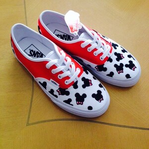 Puede incluir: Un par de zapatos de lona blancos con dise&ntilde;os de Mickey Mouse rojos y negros. Los zapatos tienen cordones negros y la palabra "VANS" est&aacute; impresa en el lateral.