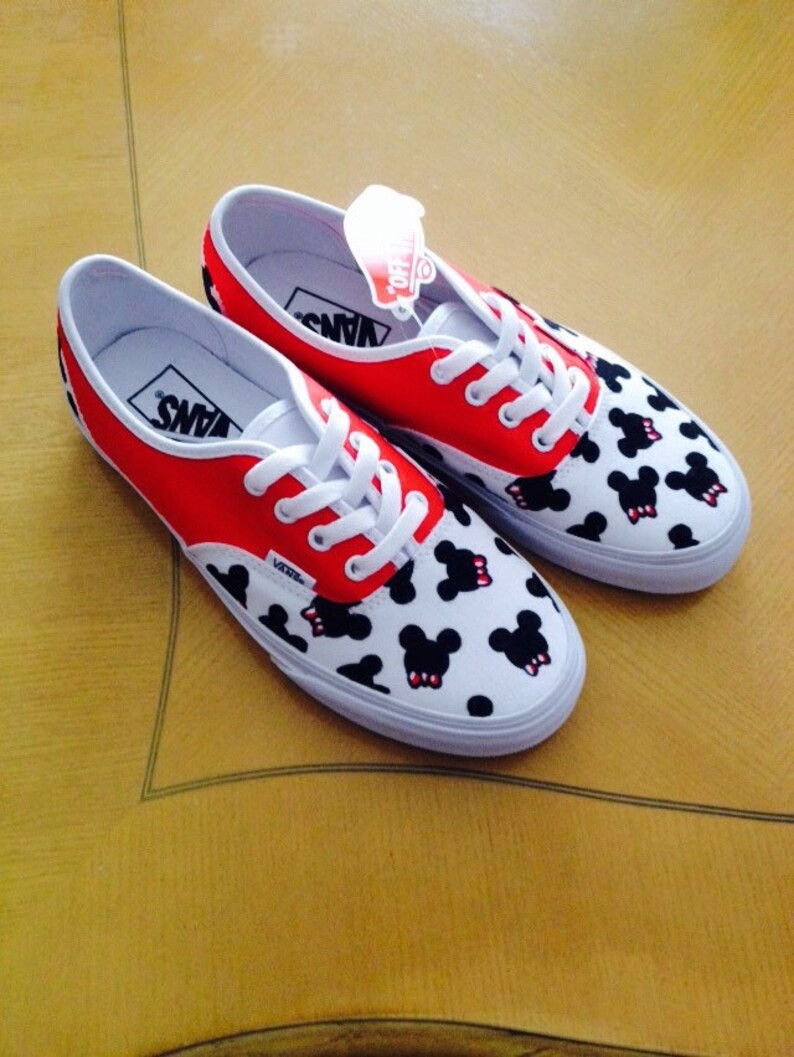 Puede incluir: Un par de zapatos de lona blancos con dise&ntilde;os de Mickey Mouse rojos y negros. Los zapatos tienen cordones negros y la palabra "VANS" est&aacute; impresa en el lateral.