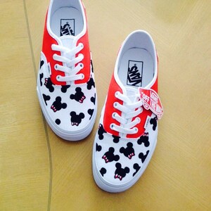 Puede incluir: Un par de zapatillas Vans rojas y blancas con estampado de Mickey Mouse. Las zapatillas tienen cordones blancos y un logotipo negro de Vans en el lateral.