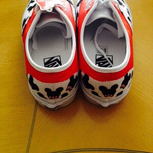 Puede incluir: Un par de zapatos de lona blancos con ribetes rojos y siluetas negras de Mickey Mouse. Los zapatos tienen cordones blancos y un logotipo negro de Vans en el lateral.