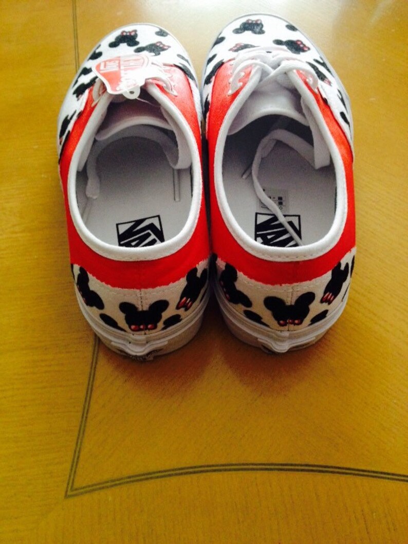 Puede incluir: Un par de zapatos de lona blancos con ribetes rojos y siluetas negras de Mickey Mouse. Los zapatos tienen cordones blancos y un logotipo negro de Vans en el lateral.