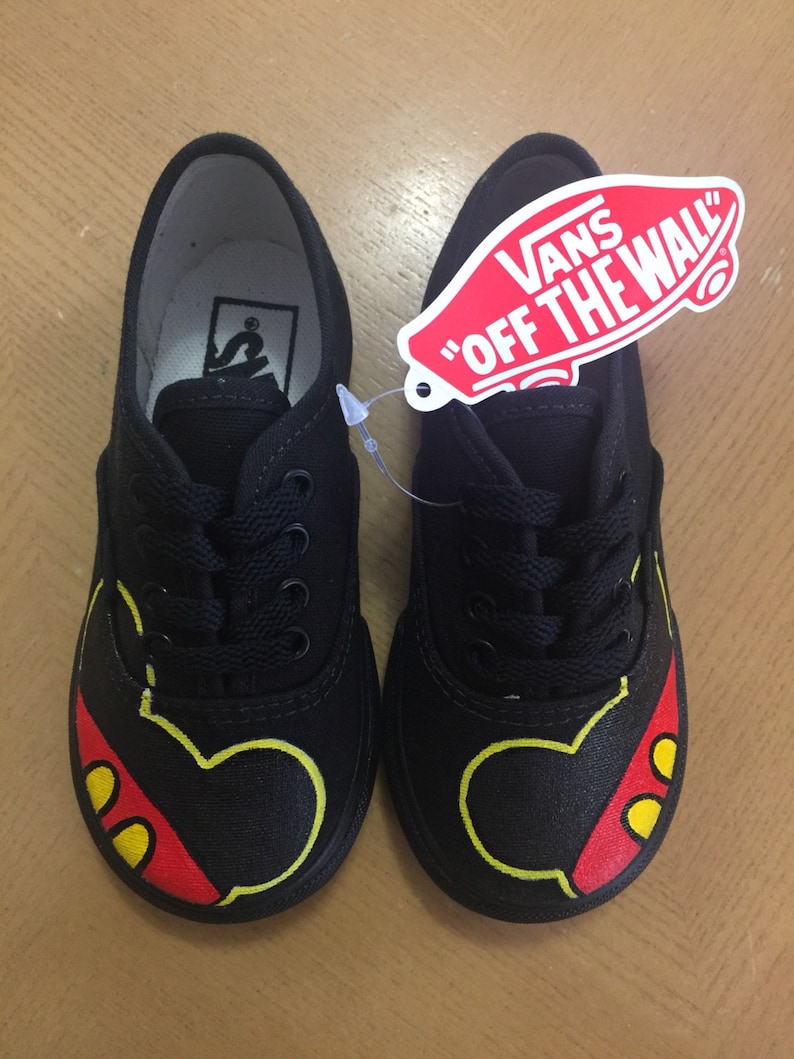 May include: Black canvas Vans shoes with a Mickey Mouse design painted on them. The shoes have yellow outlines and red and yellow accents. The Vans logo is on the side of the shoes.
