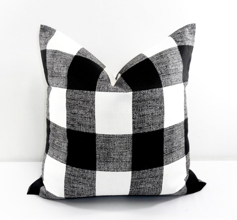 May include: Square decorative pillow with a black and white buffalo check pattern. The pillow has a textured appearance and a classic, farmhouse style. The black and white squares are evenly sized and create a bold visual statement. The pillow is a neutral color and can be used in any room.