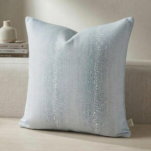 以下が含まれることがあります： ライトブルーと白の正方形の装飾用クッション。 クッションは、斑点模様のテクスチャード加工の外観をしています。 ホームデコレーションに適した、明るい色の表面に配置されています。