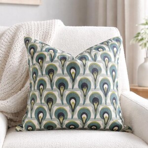 Pu&ograve; includere: Un cuscino decorativo con un motivo a gocce verde acqua, blu navy e verde su uno sfondo a righe grigio chiaro. Il cuscino &egrave; posto su una poltrona bianca, con una coperta color crema drappeggiata sulla sedia.
