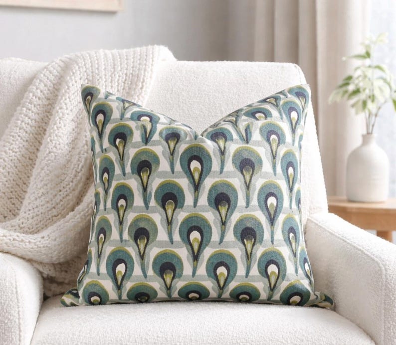 Pu&ograve; includere: Un cuscino decorativo con un motivo a gocce verde acqua, blu navy e verde su uno sfondo a righe grigio chiaro. Il cuscino &egrave; posto su una poltrona bianca, con una coperta color crema drappeggiata sulla sedia.