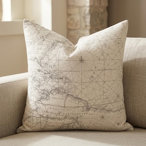 May include: A decorative throw pillow with a vintage map design. The pillow features a detailed, faded gray map print on a cream-colored background. The map includes cartographic details and compass roses, adding a touch of classic decor.