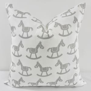 May include: White decorative pillow with a repeating pattern of gray rocking horses. The pillow has a square shape and is made of a soft fabric. The rocking horse design adds a playful touch to the pillow.