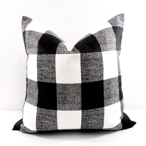 May include: Square decorative pillow with a black and white buffalo check pattern. The pillow has a textured appearance and a classic, farmhouse style. The pillow is a square shape and is suitable for a couch or bed.