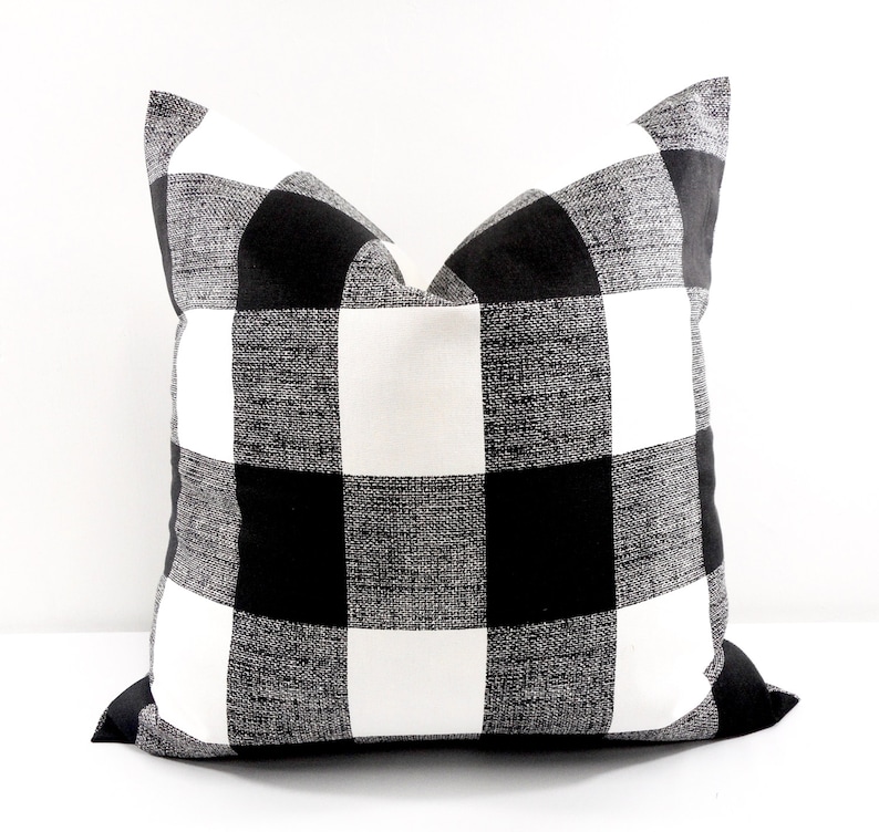 May include: Square decorative pillow with a black and white buffalo check pattern. The pillow has a textured appearance and a classic, farmhouse style. The pillow is a square shape and is suitable for a couch or bed.