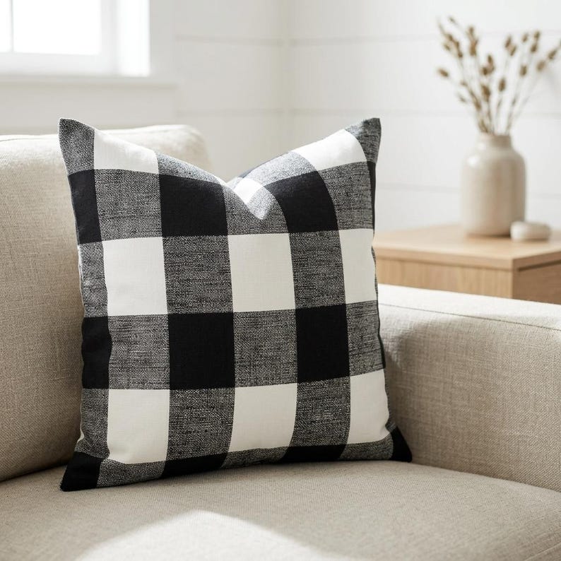 May include: A black and white buffalo check pattern pillow. The pillow is square and rests on a neutral-colored sofa. The pillow's design features large, evenly spaced squares of black and white. The pillow is a decorative home accent.
