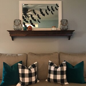 May include: A living room decorated for Halloween. A dark wooden shelf holds decorative items, including two glass lanterns, a small pumpkin, and a candle. Black bat cutouts are attached to the mirror above. The couch has green and black and white checkered pillows.